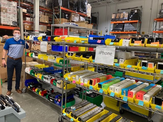 Man standing in a warehouse beside shelves filled with sorted mobile devices labeled by model.